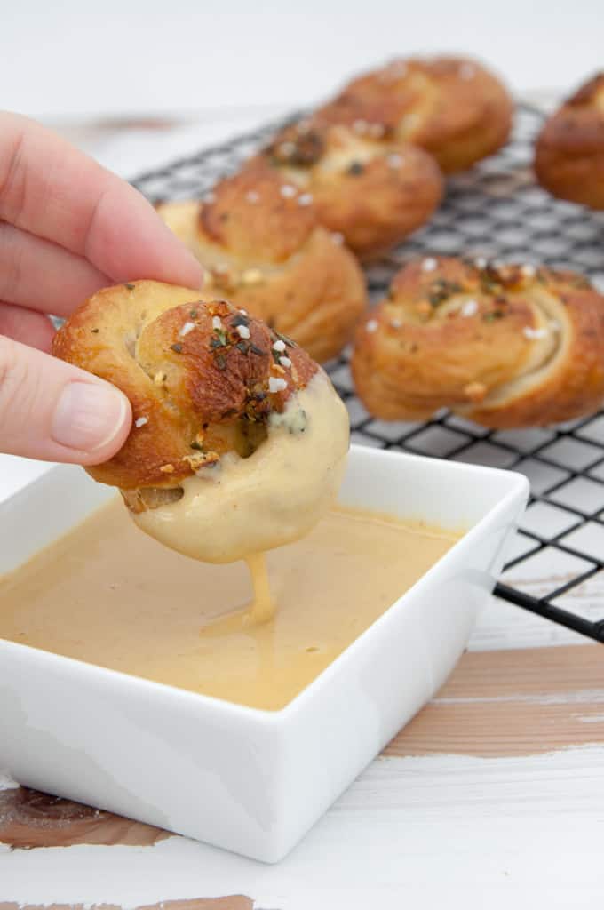 a hand dipping a Vegan Pretzel Garlic Knot in a white bowl with vegan cheese sauce with more garlic knots in the background