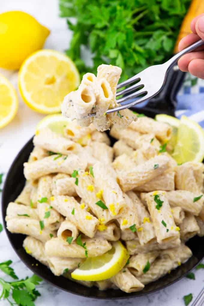 a hand holding a fork with three rigatoni over a pan of lemon pasta with parsley and lemons in the background