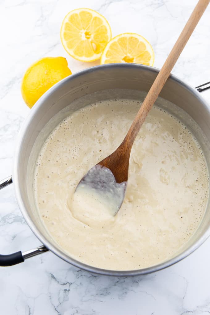 creamy lemon sauce in a pot with a wooden spoon on a marble countertop with two lemons in the background
