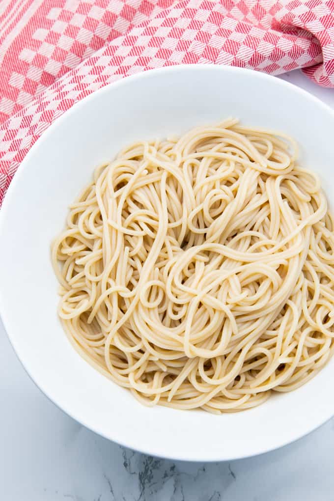 cooked spaghetti in a white bowl on a marble countertop with a red dishtowel in the background
