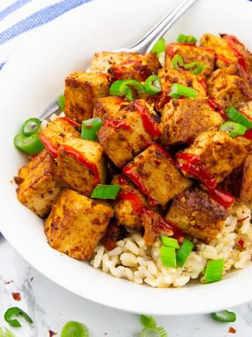 Marinated Tofu over brown rice sprinkled with chopped green onions in a white plate on a marble countertop