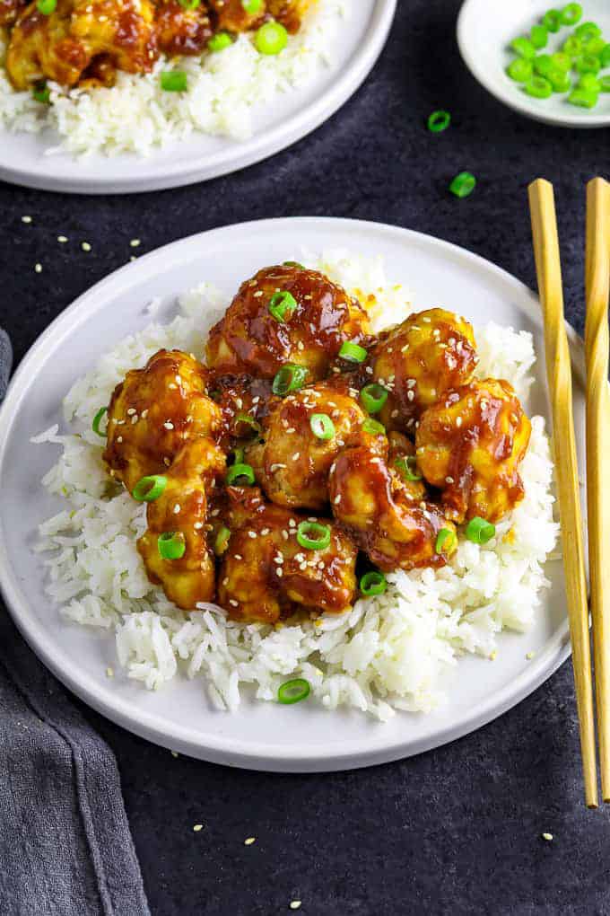 Sticky Sesame Cauliflower served over white rice on a white plate with chop sticks and another plate in the background