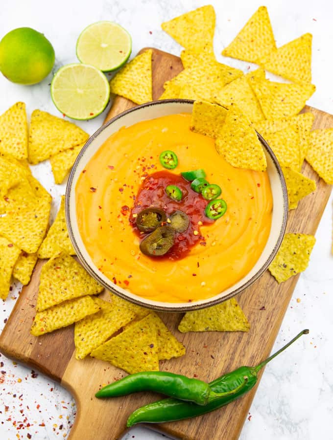 Vegan Nacho Cheese in a bowl on a wooden board with nachos, two jalapeños, and two limes on the side