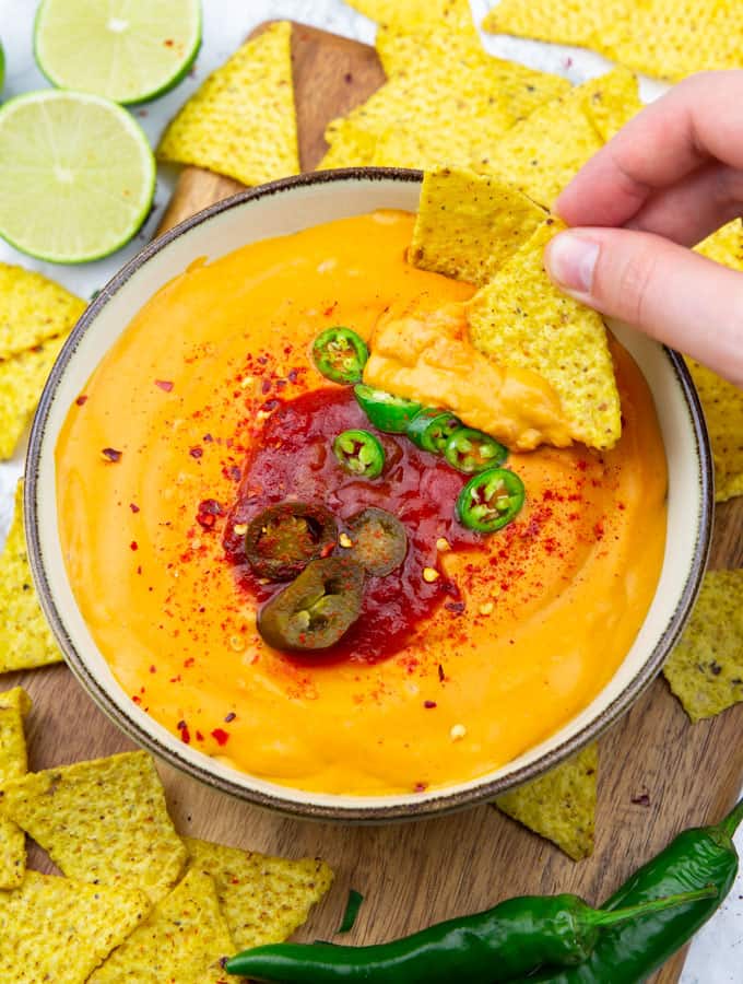 a bowl with vegan nacho cheese on a wooden board with nachos, two lime halves, and jalapeños in the background and a hand dipping a nacho into the cheese sauce 