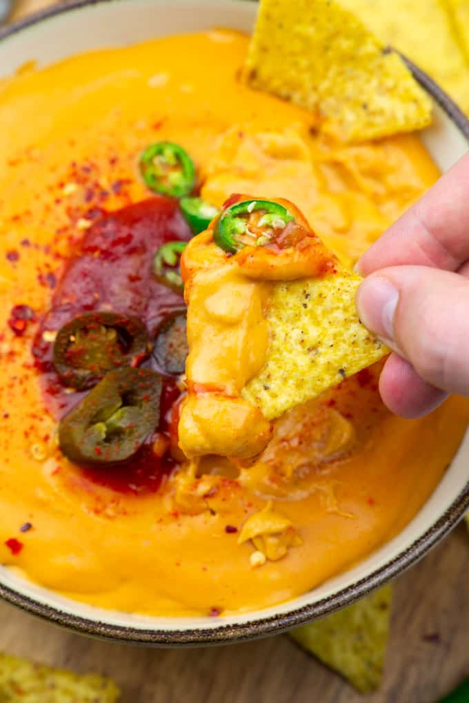 a hand dipping a nacho into a bowl with vegan nacho cheese with salsa and jalapeños on top