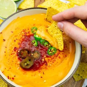a bowl with vegan nacho cheese on a wooden board with nachos, two lime halves, and jalapeños in the background and a hand dipping a nacho into the cheese sauce