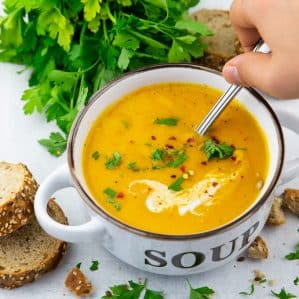 Vegan Pumpkin Soup in a white and brown bowl with a hand holding a spoon and a bunch of parsley in the background