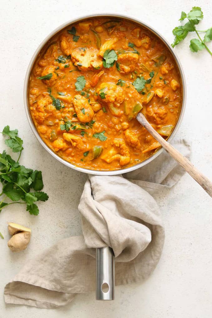 a pan of Cauliflower and Chickpea Curry with a wooden spoon on a beige countertop with fresh cilantro and ginger on the side 