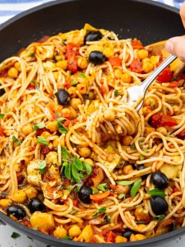 vegan spaghetti with chickpeas, olives, and artichokes hearts in a black pan with a wooden spoon on a marble countertop
