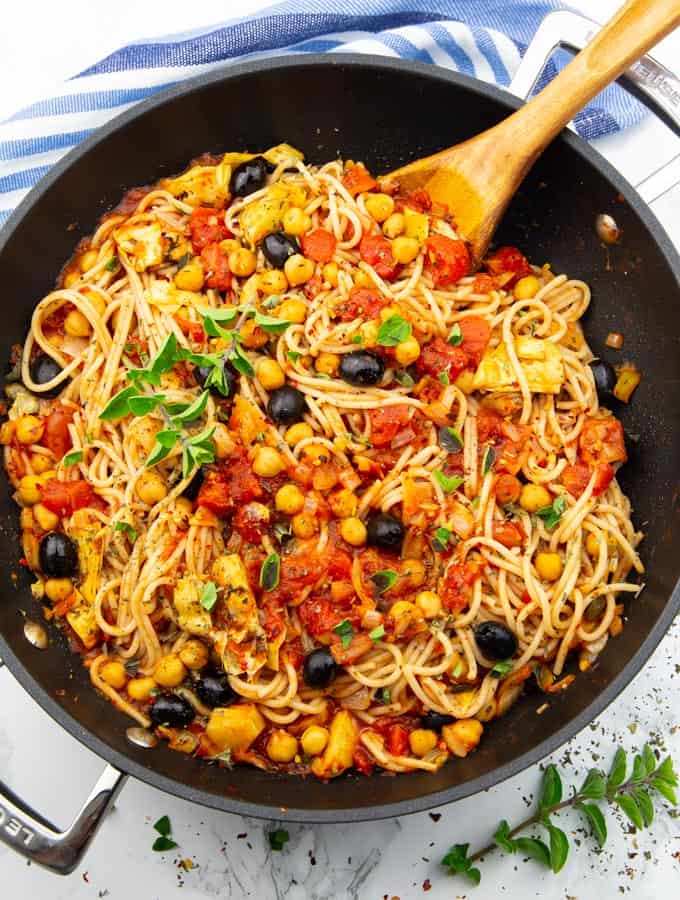 vegan spaghetti with chickpeas, olives, and artichokes hearts in a black pan with a wooden spoon on a marble countertop 