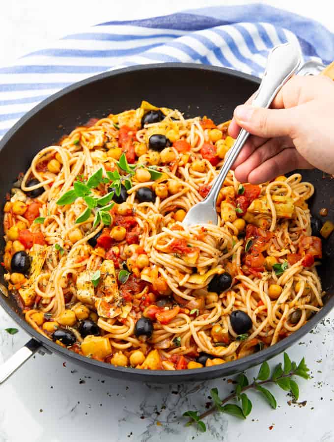 Vegan Spaghetti with tomato sauce, chickpeas, and olives in a black pan on a marble countertop with a hand rolling up some of the spaghetti with a fork