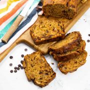 five vegan zucchini bread slices on a marble counter top with more zucchini bread on a wooden board in the background and chocolate chips on the side