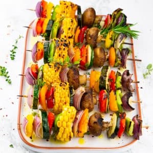 six grilled vegetable kabobs on a white plate on a marble countertop sprinkled with fresh herbs