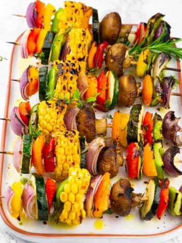 six grilled vegetable kabobs on a white plate on a marble countertop sprinkled with fresh herbs