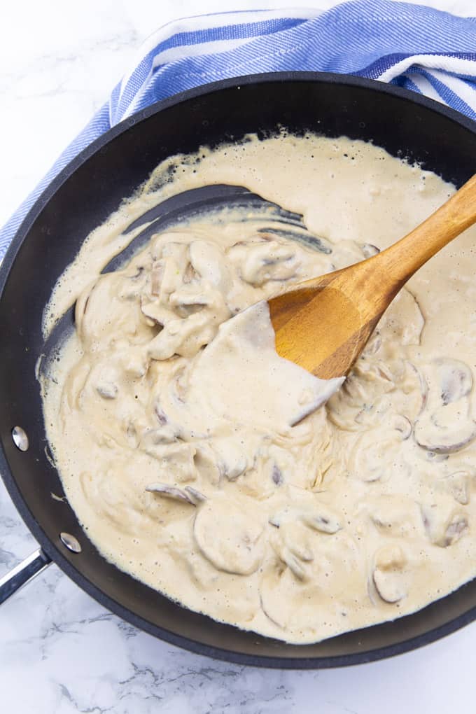 a creamy cashew-based sauce with mushrooms in a black pan with a wooden spoon on a marble countertop