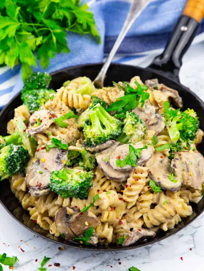 Broccoli Pasta in a black pan with a fork on a marble countertop with a with a blue tablecloth and fresh parsley in the background