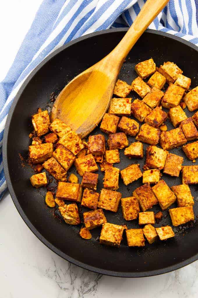 marinated tofu cubes in a black pan with a wooden spoon on a marble countertop 