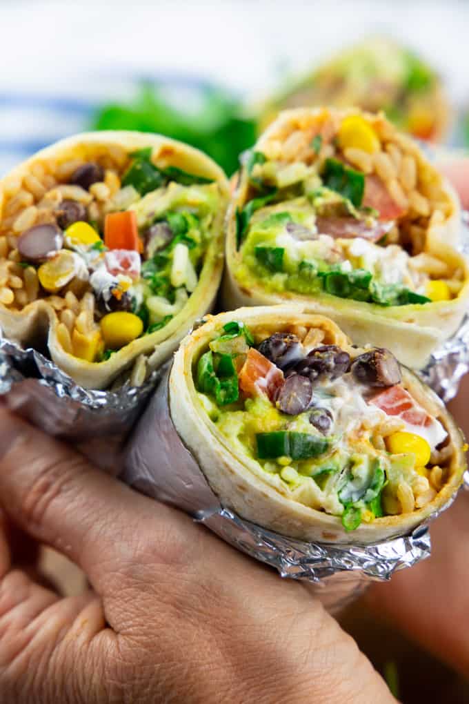 a hand holding three vegan burritos wrapped in aluminium foil 