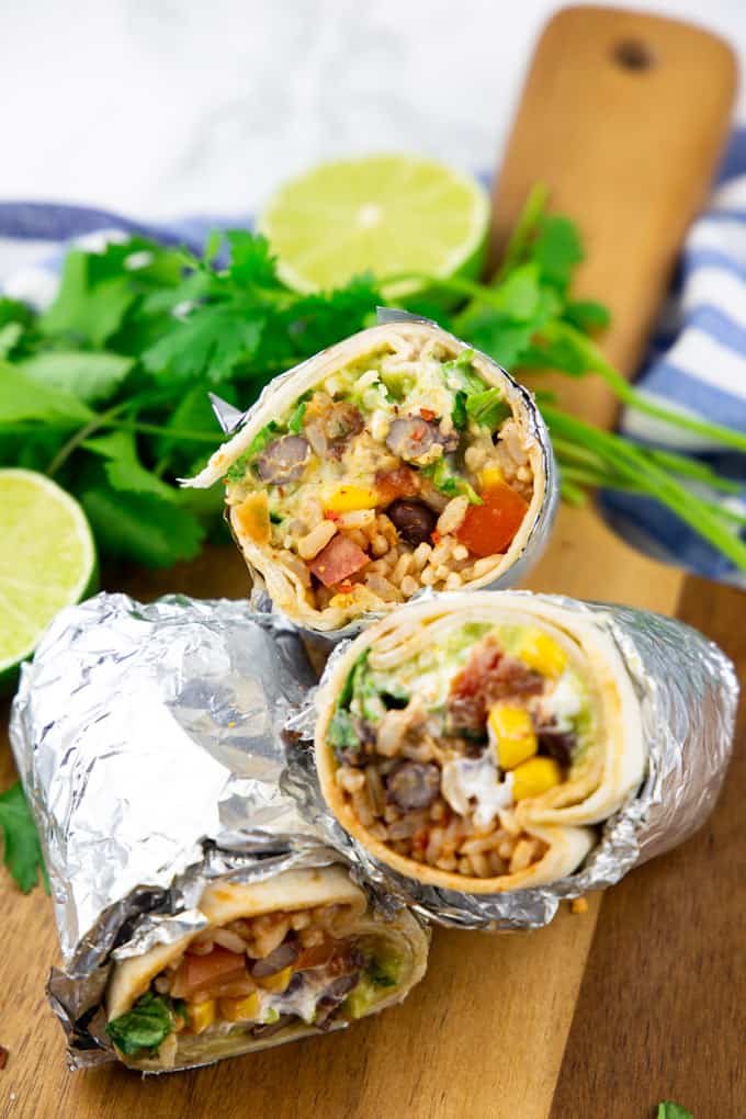 three vegan burritos wrapped in aluminium foil on a wooden board with limes and cilantro in the background