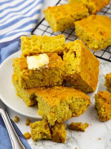 a stack of vegan cornbread squares on a white plate with more cornbread in the background on a cooling rack and a butter knife on the side
