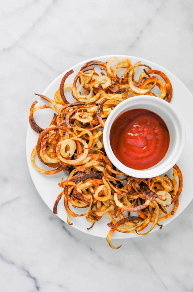 curly fries on a white plate with a bowl of ketchup on the side on a marble countertop 