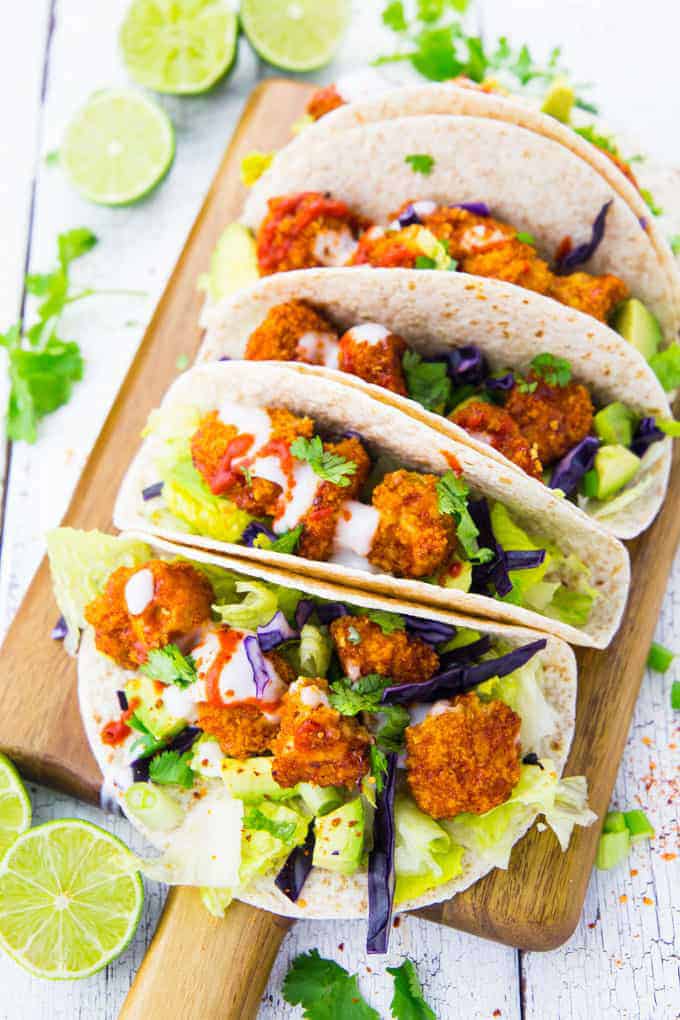 four vegan cauliflower tacos on a wooden board with lime halves and fresh cilantro on the side 