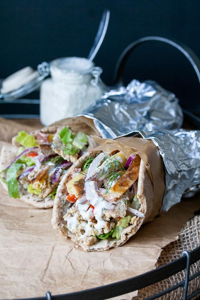 two Tempeh Gyros wraps wrapped in aluminium foil on parchment paper with a small jar of sauce in the background