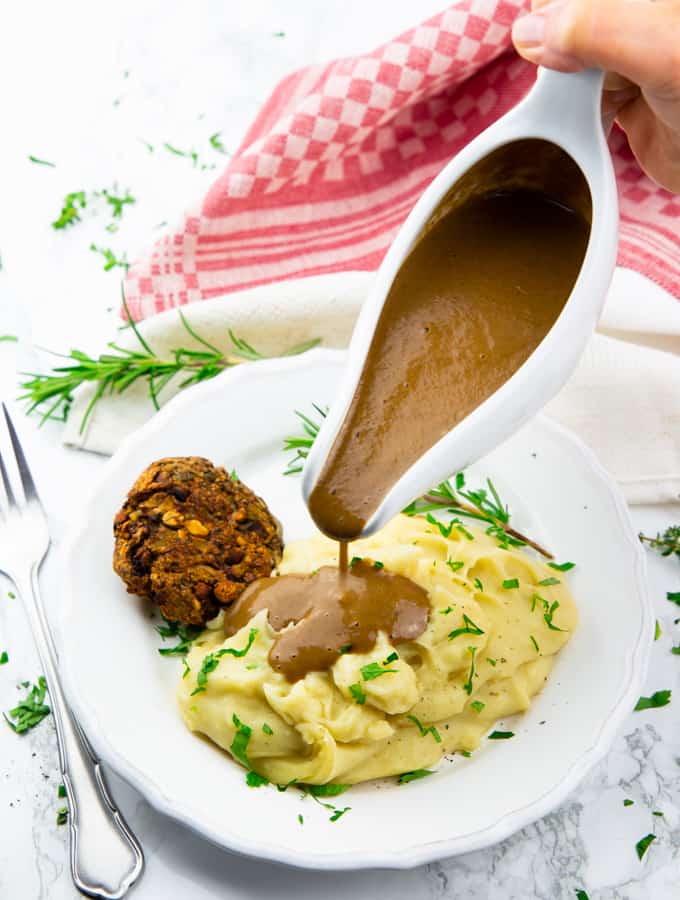 a hand pouring vegan gravy out of a sauce boat over mashed potatoes on a white plate with fresh herbs and a fork on the side 