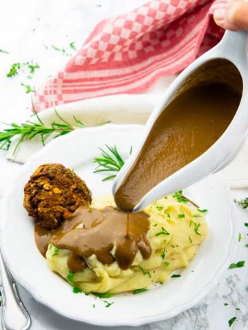 a hand pouring vegan gravy out of a sauce boat over mashed potatoes on a white plate with fresh herbs and a fork on the side