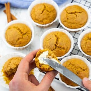 a hand spreading butter on a pumpkin muffin with a knife with more vegan pumpkin muffins in the background