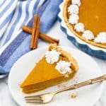 a slice of vegan pumpkin pie on a white plate with a fork on the side and more pumpkin pie and two cinnamon sticks in the background