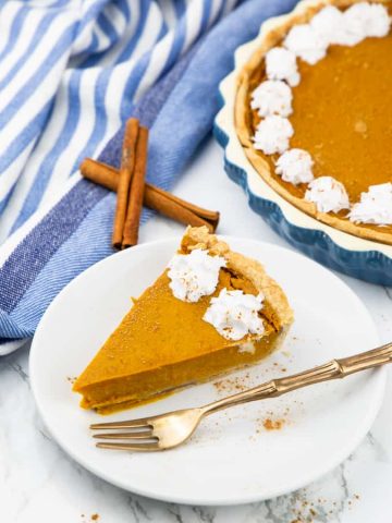a slice of vegan pumpkin pie on a white plate with a fork on the side and more pumpkin pie and two cinnamon sticks in the background