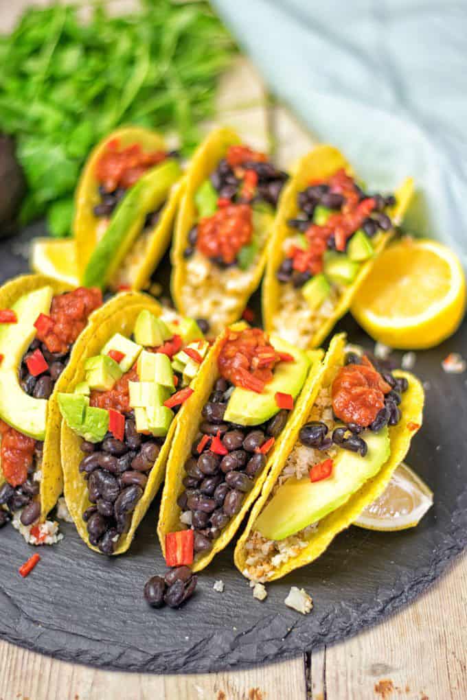 seven Mexican Cauliflower Rice Tacos on a black board with lemon halves on the side and cilantro in the background