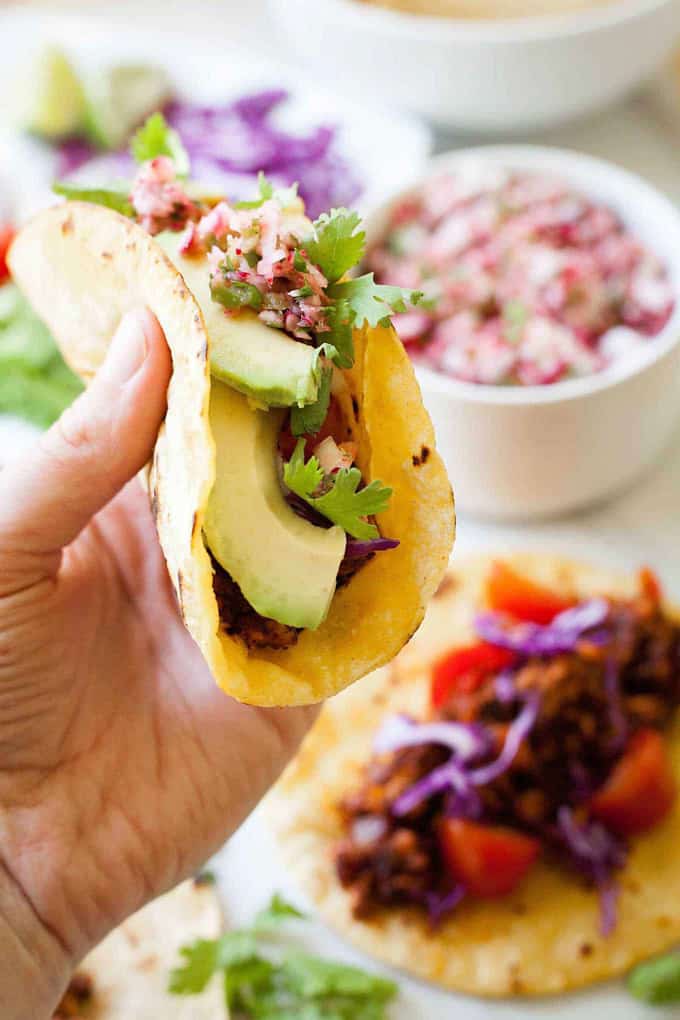 a hand holding a vegan taco with tempeh and avocado with more tacos and bowls with filling in the background