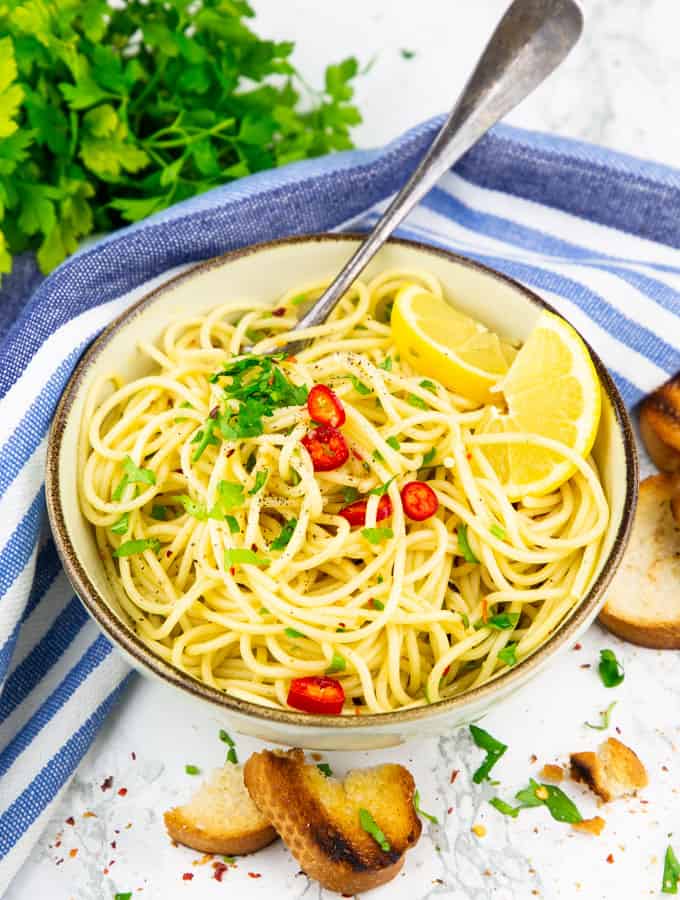 spaghetti aglio e olio in a beige and brown bowl with lemon slices on top on a marble countertop and roasted bread and a bunch of parsley on the side