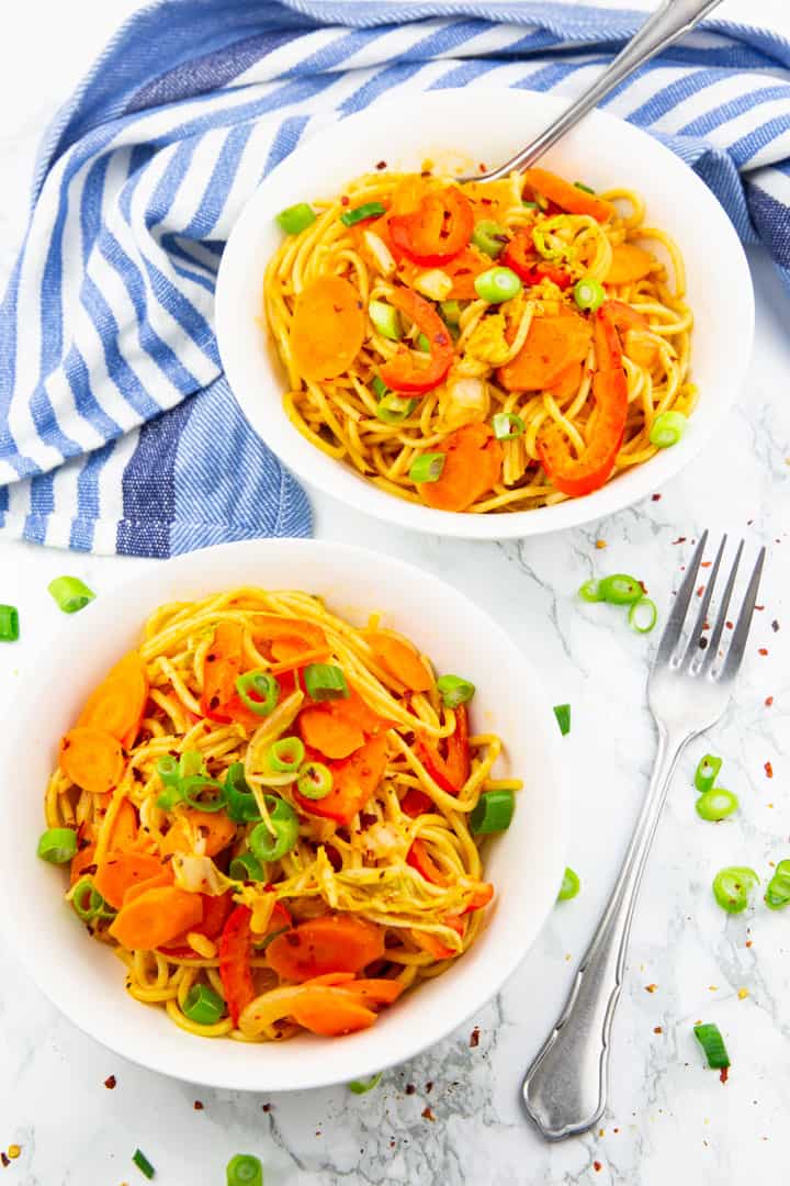 Asian spaghetti with red bell pepper and carrots in two white bowls on a marble countertop with a fork and chopped green onions on the side