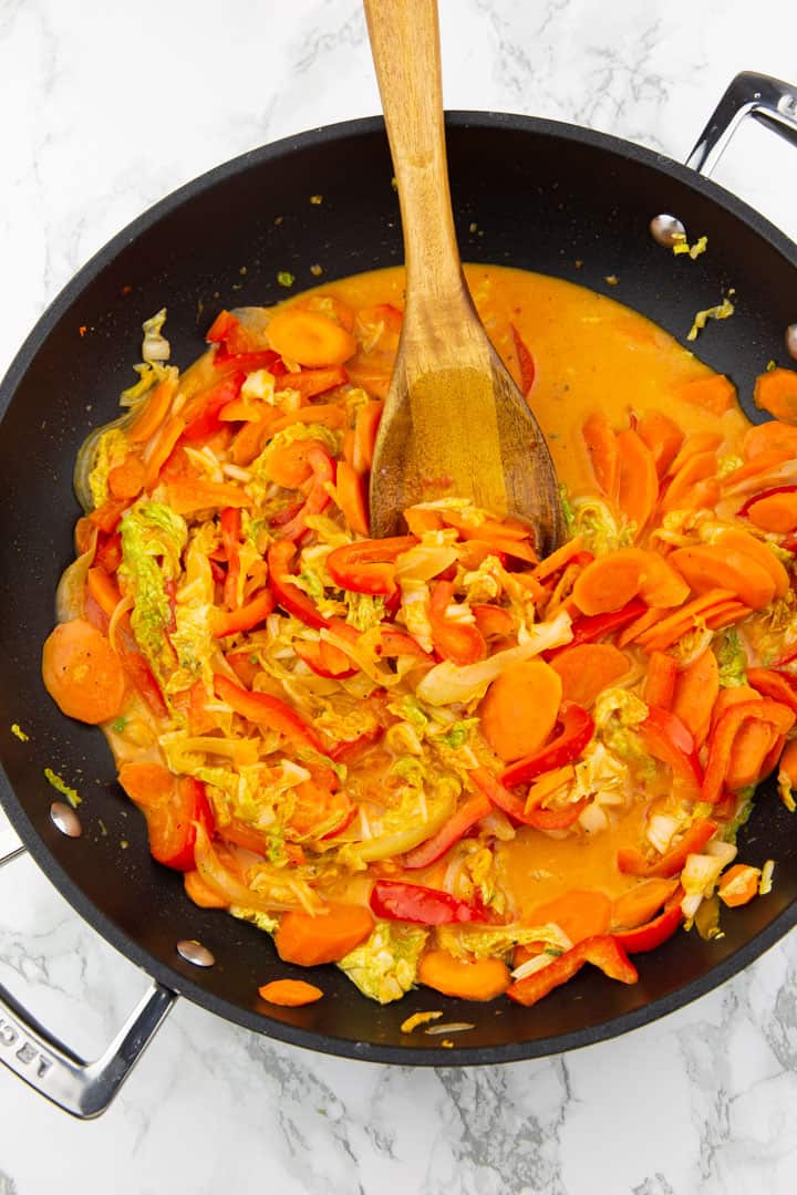 red curry with bell pepper, carrots, and Chinese cabbage in a black pan with a wooden spoon on a marble countertop 