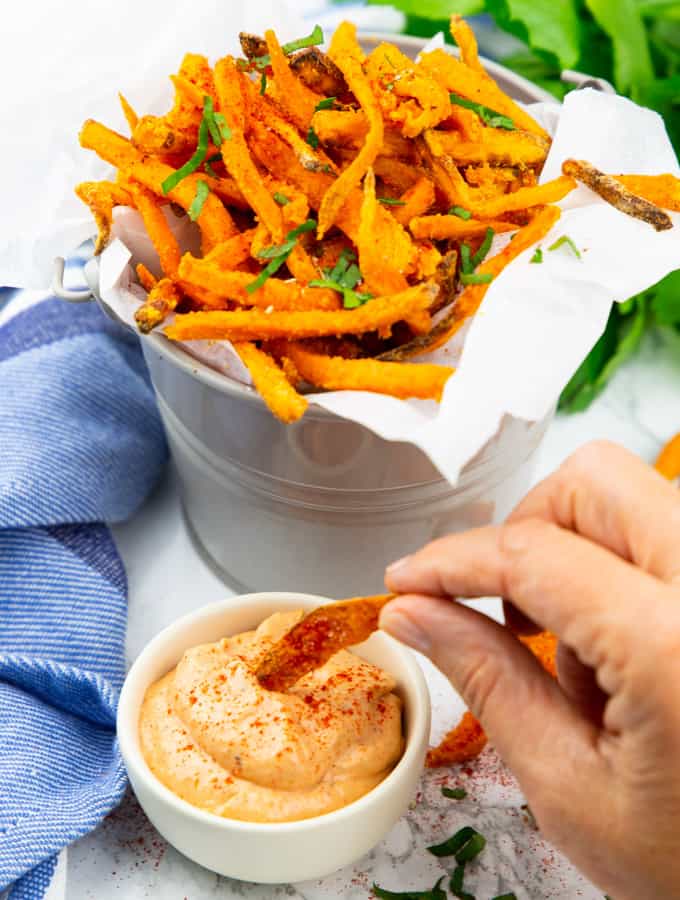 a hand dipping sweet potato fries into a small bowl of sweet potato fries dipping sauce with more fries in the background