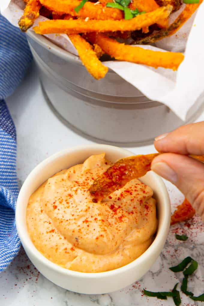 Sweet Potato Fries Dipping Sauce in a white bowl on a marble countertop with a hand dipping a sweet potato fry into the dip 