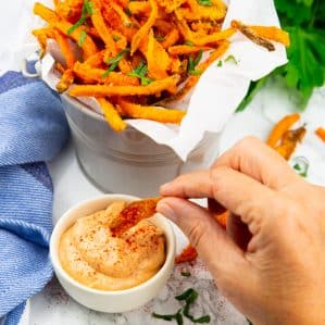 a hand dipping sweet potato fries into a small bowl of Cajun-spiced sriracha mayonnaise with more fries in the background