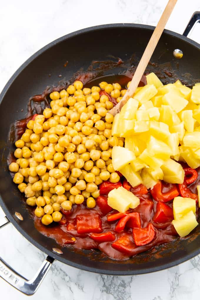 chickpeas, pineapple cubes, and red bell pepper with a sweet and sour sauce in a black pan with a wooden spoon on a marble countertop 