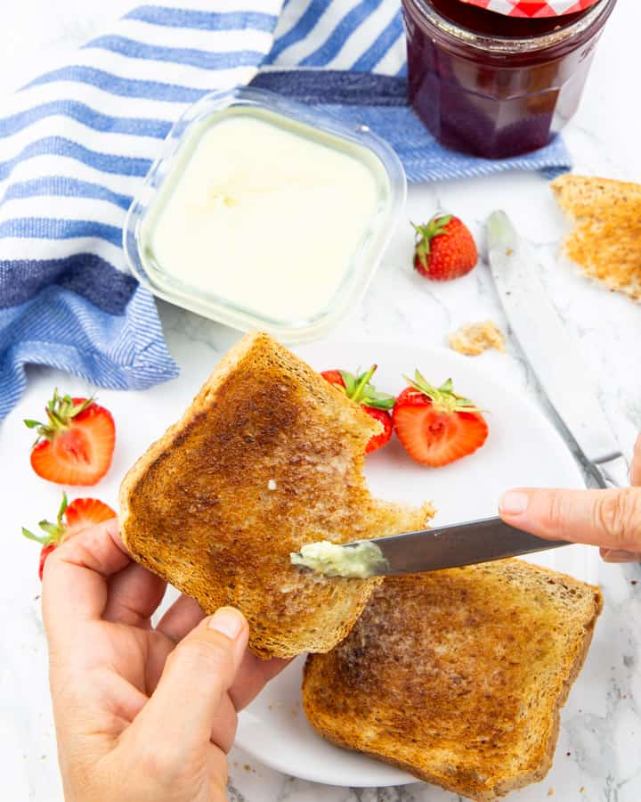 a hand holding a toast and spreading vegan butter on with strawberries and jam in the background