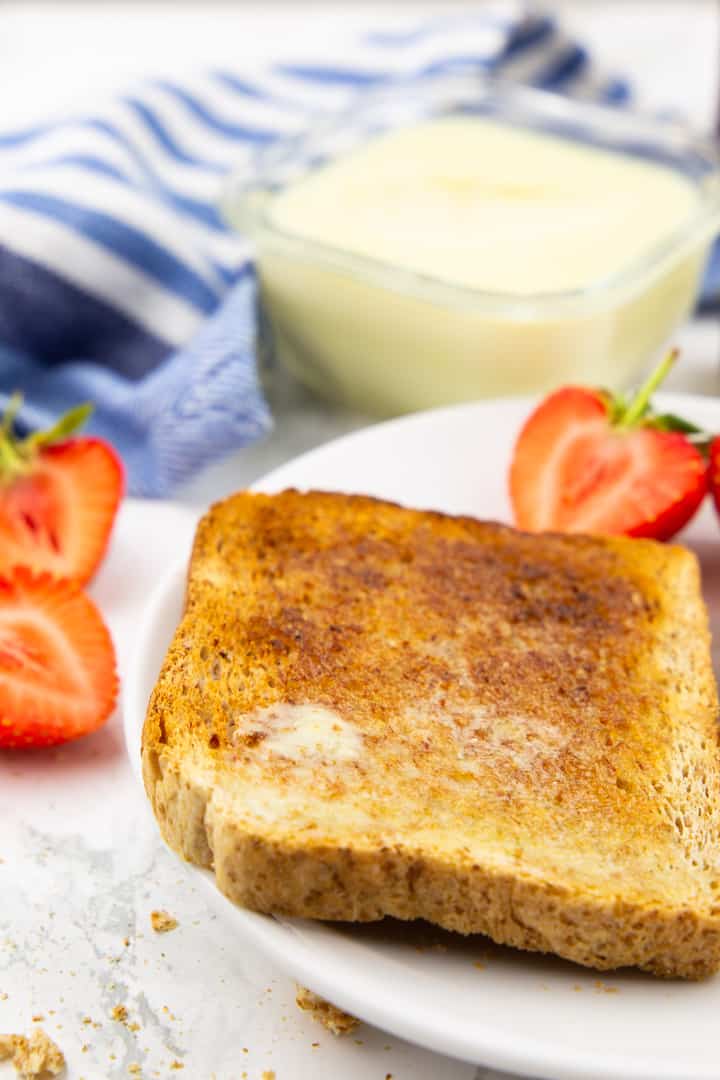 a slice of toast on a white plate with a small bowl of vegan butter in the background and strawberries on the side 