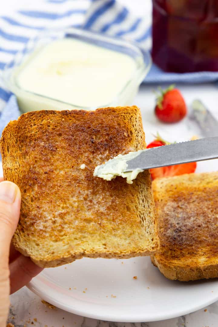 a hand holding a slice of toast and spreading vegan butter on it with a butter knife 