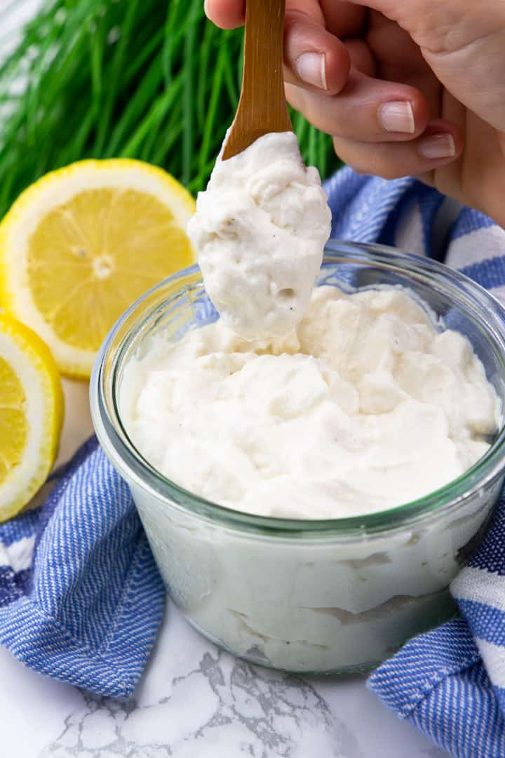 Vegan Mayonnaise in a glas jar with a hand holding a little wooden spoon with mayonnaise with chives and a lemon in the background