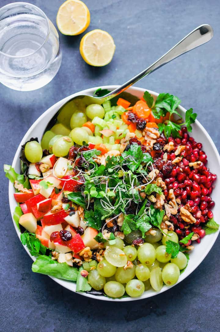 a detox salad with pomegranate, grapes, and lettuce on a dark countertop with a glas of water and two lemon halves on the side