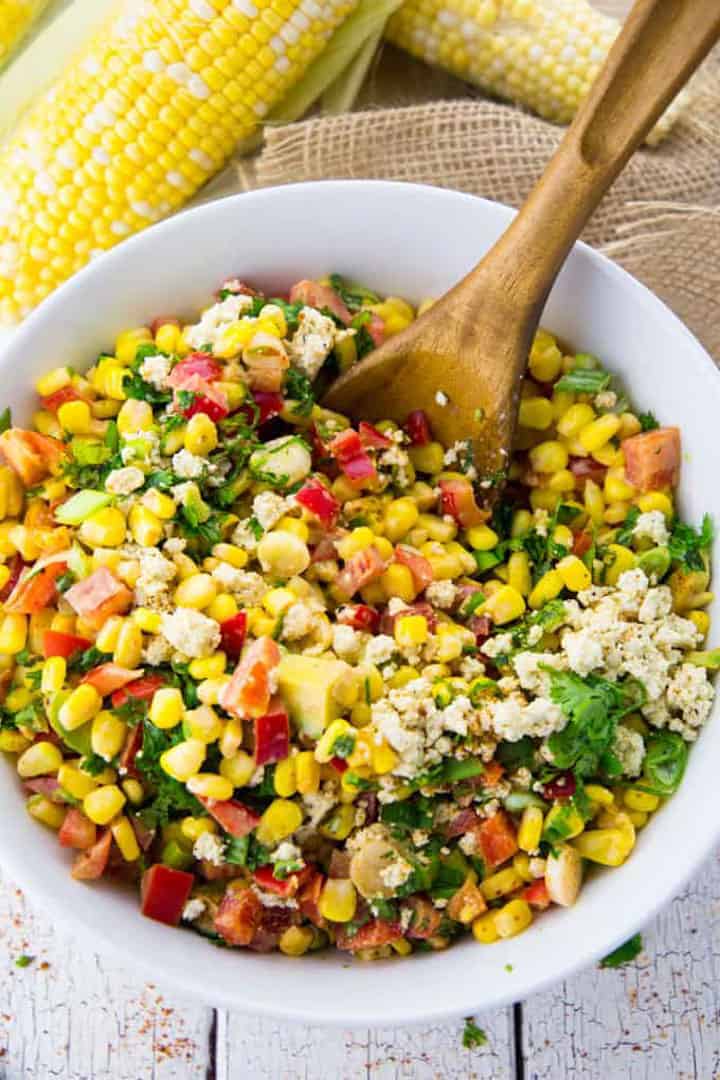 vegan corn salad in a white bowl on a white wooden countertop with a wooden spoon and two corn cobs in the background