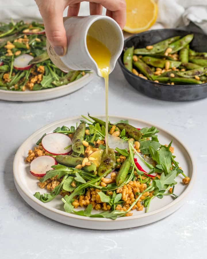 Vegan Snap Pea Arugula Farro Salad on two white plates with a hand pouring the dressing over the salad 