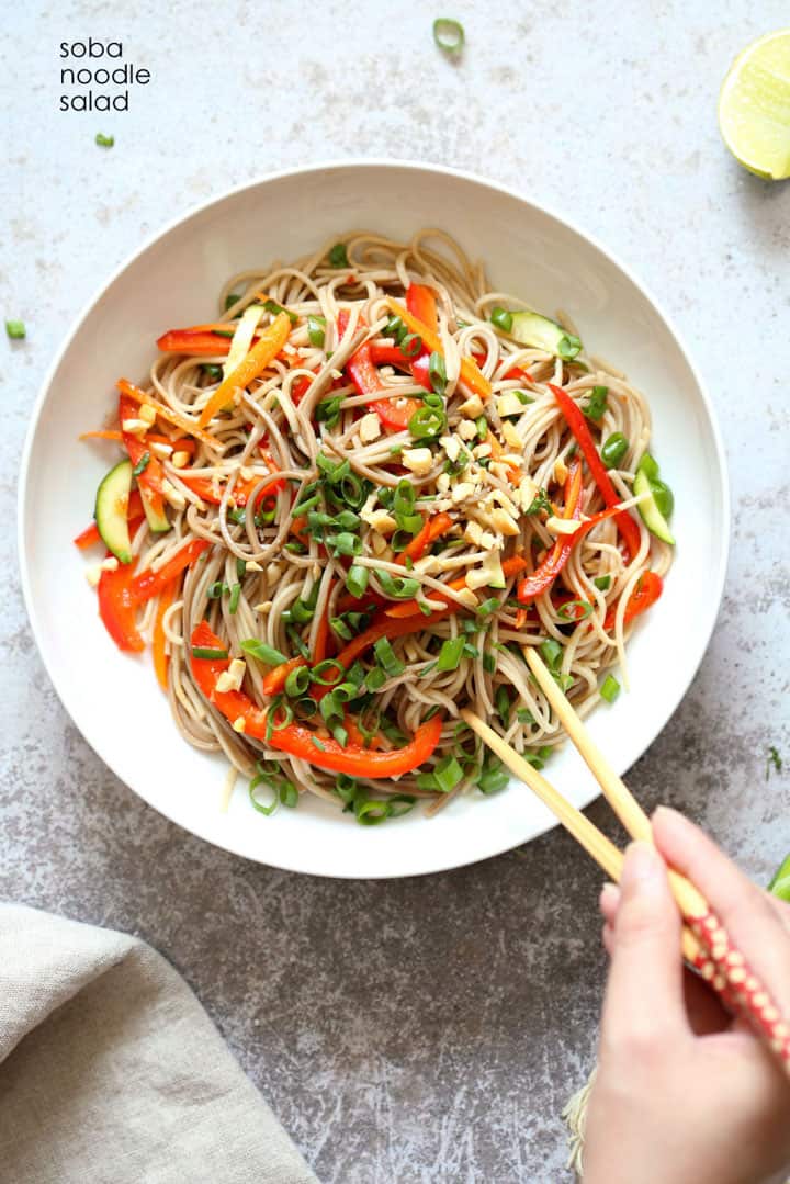 cold soba noodle salad in a white bowl on a concrete countertop with a hand picking up some of the noodles with two chopsticks 