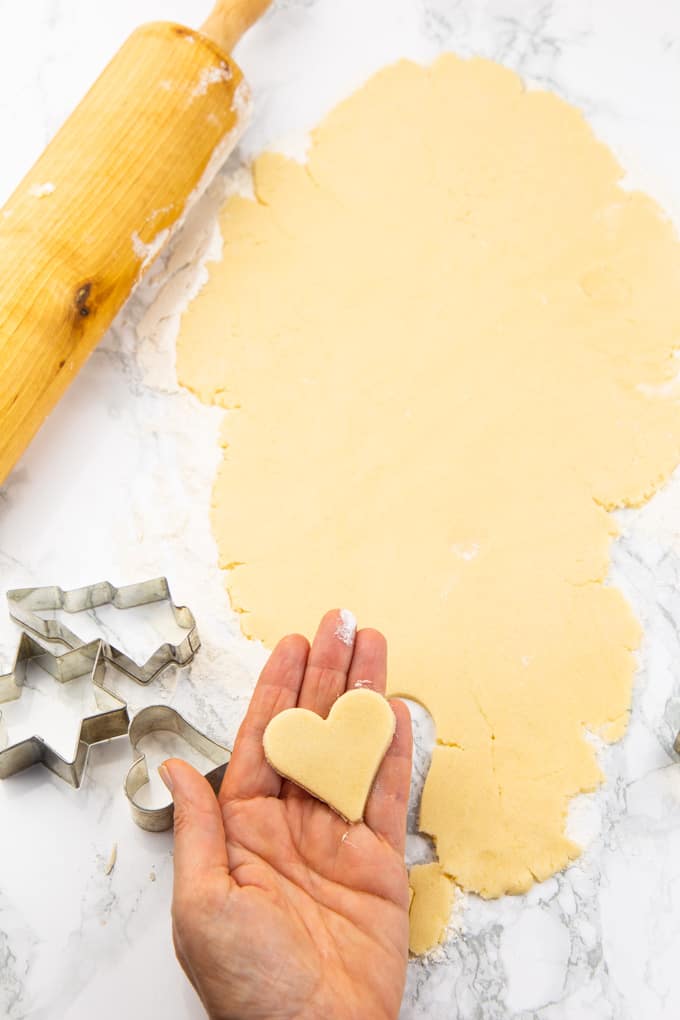 a hand holding an unbaked heart-shaped sugar cookie with cookie dough and a rolling pin in the background 